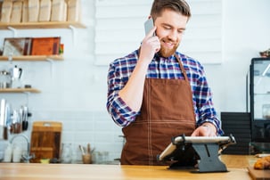 Handsome smiling barista with beard taking order on cell phone and using tablet in cafeteria-1